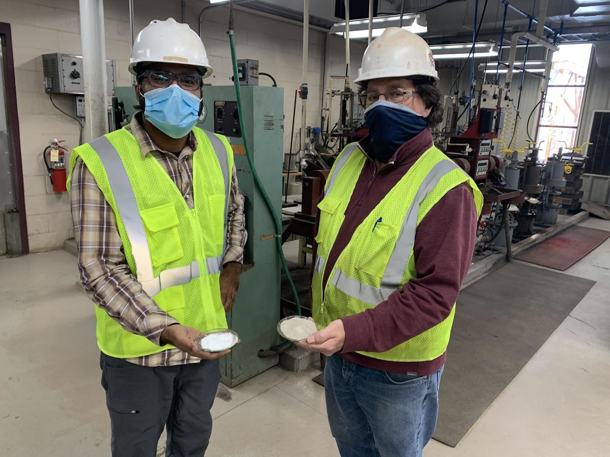 Two men wearing safety PPE holding small dish each with powdery material.