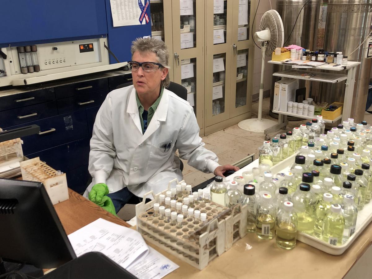 A woman in white lab coat and safety glasses sits behind a desk covered with small liquid bottles.