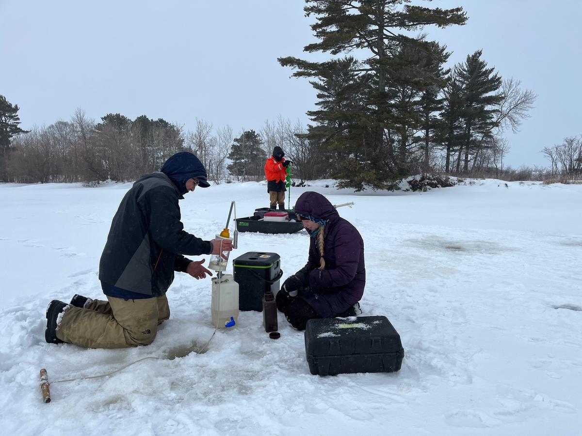 Scientists kneel and use water sampling equpment next to a small hole in lake ice.