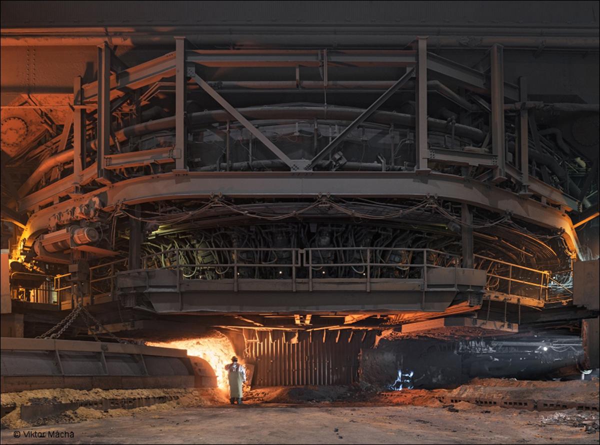 Industrial scene of large glowing furnace, person in front dwarfed by machinery.
