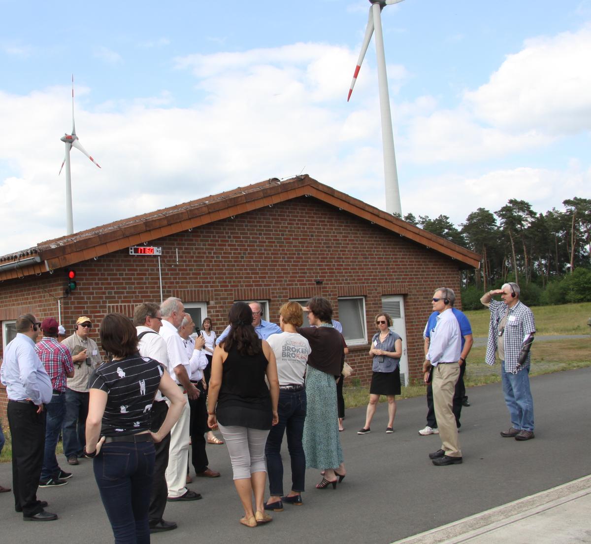 A group of people surrounding person speaking with wind turbines in background
