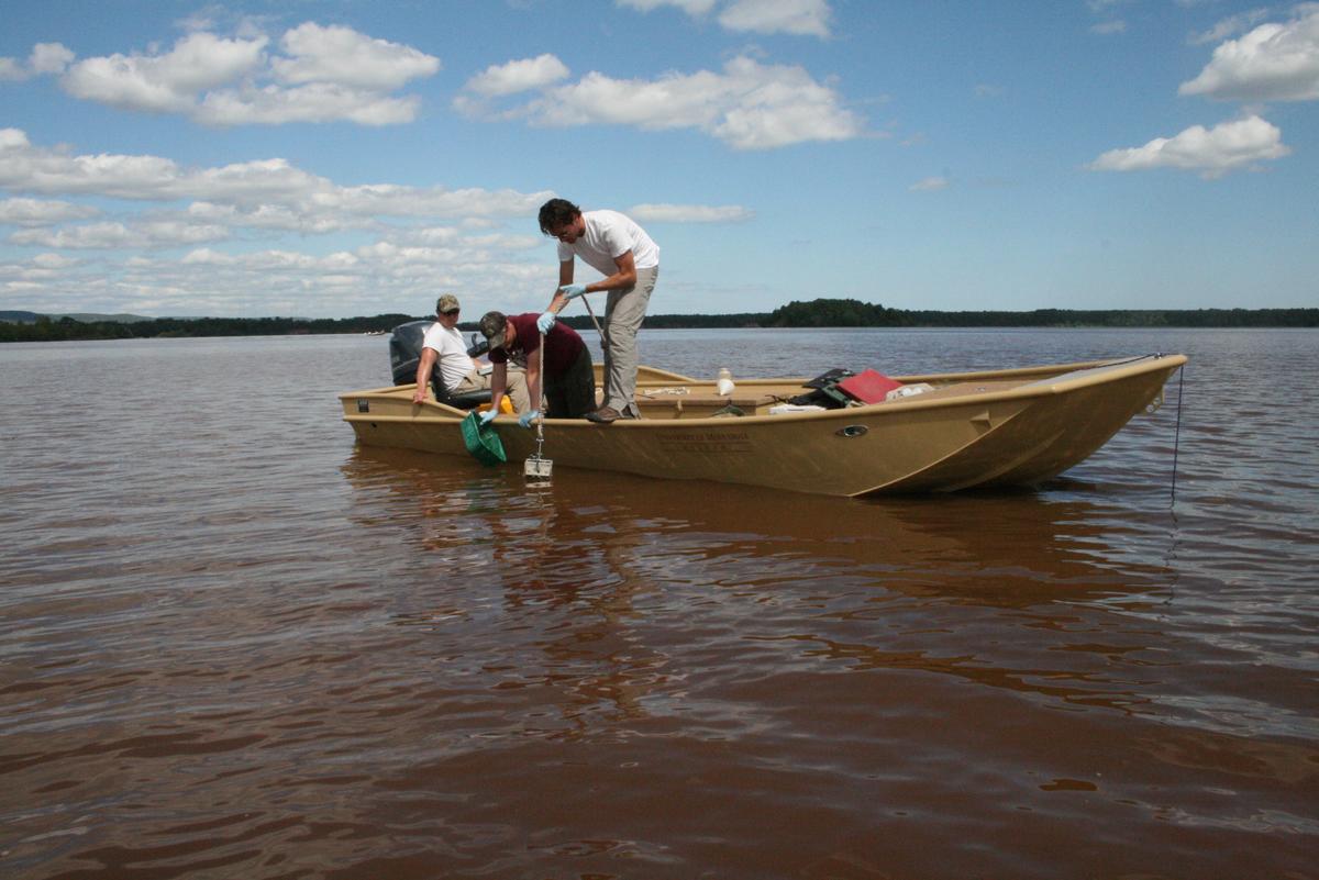 Three males in a low profile boat on a large lake, two males dropping a tool into the water.