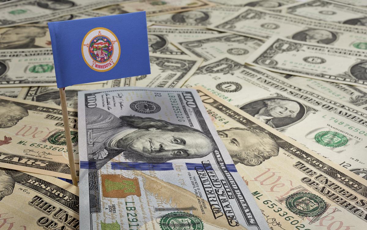 U.S. paper currency scattered with a state of Minnesota flag flying above.