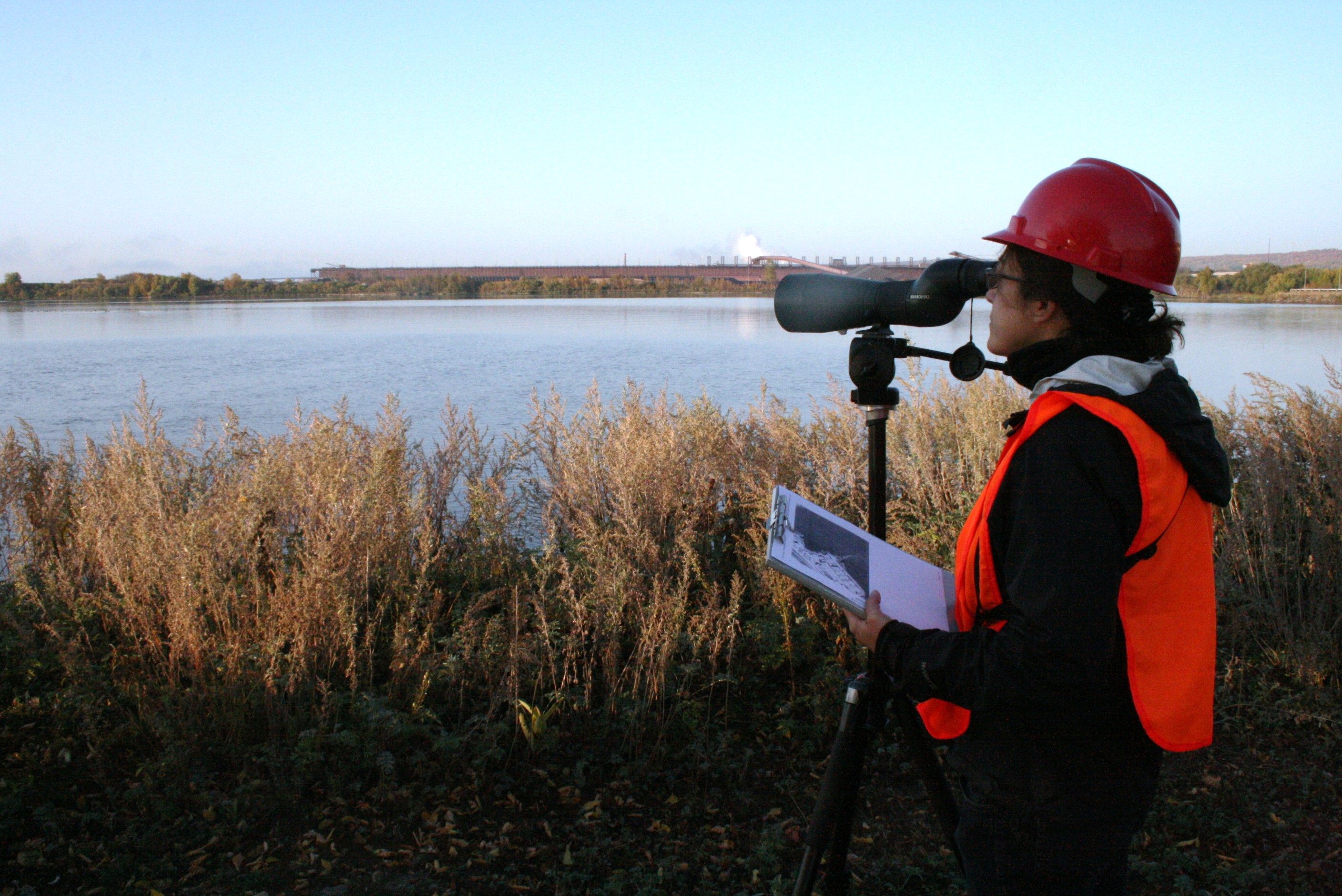 Woman in safety vest and hard hat looks through telescope at dawn by water body.